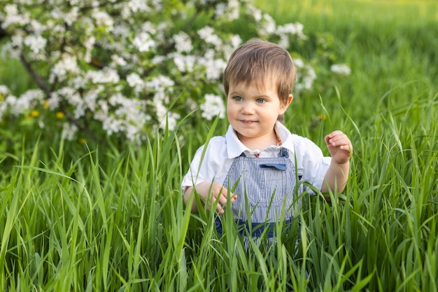 Szczęśliwe małe dziecko w modnym niebieskim kombinezonie z pięknymi niebieskimi oczami. zabawne zabawy w wysokiej zielonej trawie w kwitnącym parku pełnym zieleni na tle jabłoni.