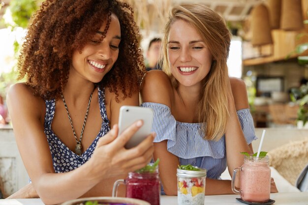 Szczęśliwe młode kobiety oglądają śmieszne filmy w internecie na smartfonie, siadają razem na tle kawiarni, jedzą smaczny deser i świeże koktajle. Koncepcja ludzi, relacji i nowoczesnych technologii.
