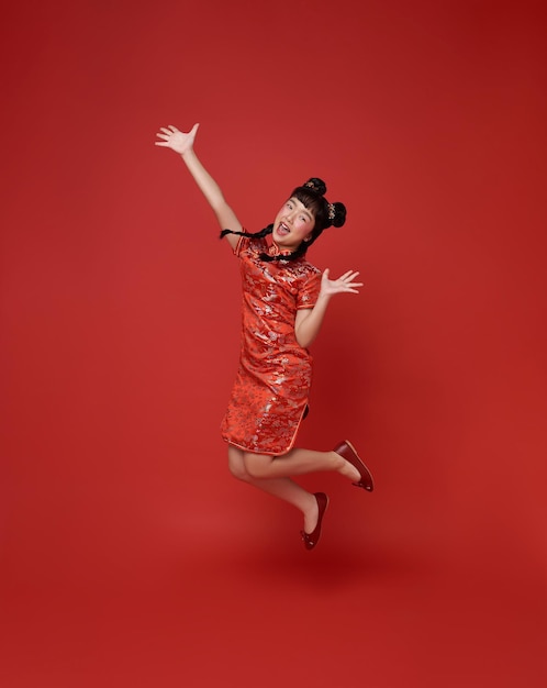 Bezpłatne zdjęcie szczęśliwego chińskiego nowego roku szczęśliwe uśmiechnięte dzieci azjatka w tradycyjnej sukience qipao skacząca świętuje izolowana na czerwonym tle