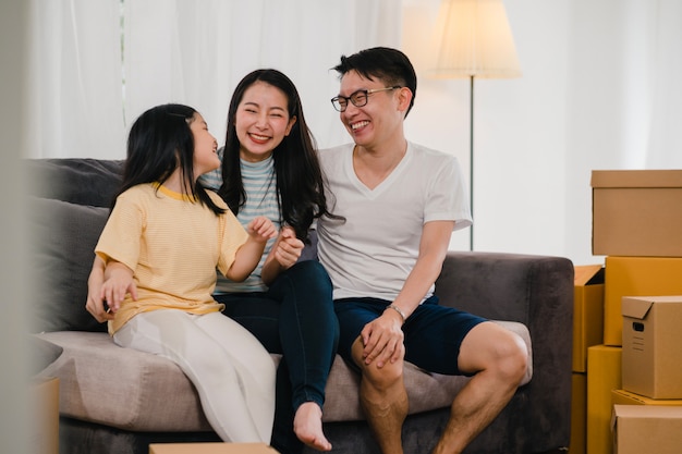 Szczęśliwi azjatyccy młodzi właściciele domów kupili nowy dom. Japońska mama, tata i córka obejmując się niecierpliwie czekają na przyszłość w nowym domu po przeprowadzce i przeprowadzce, siedząc razem na kanapie z pudełkami.