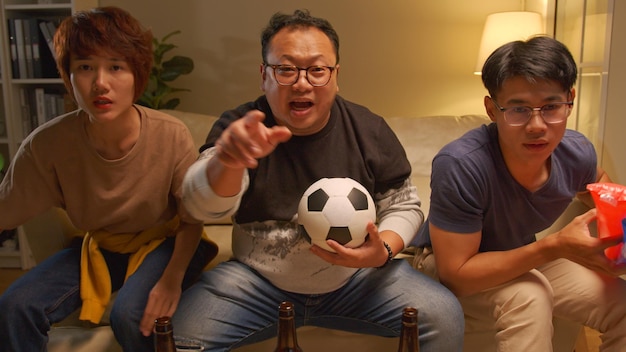 Szczęśliwi azjatyccy przyjaciele lub fani piłki nożnej oglądający piłkę nożną w telewizji i świętujący zwycięstwo w domuKoncepcja sportu i rozrywki przyjaźni