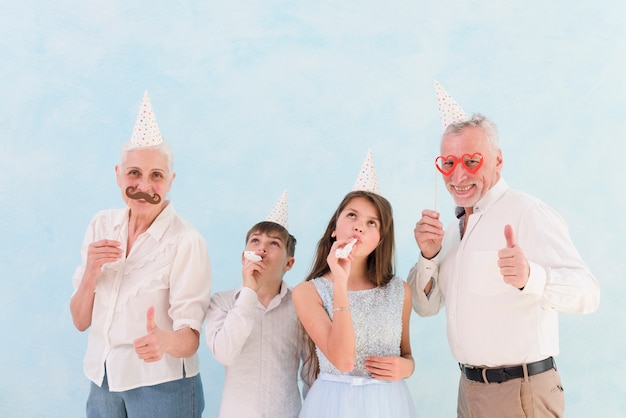 Szczęśliwi dziadkowie pokazujący papierowe rekwizyty z wnukami dmuchającymi w rogi