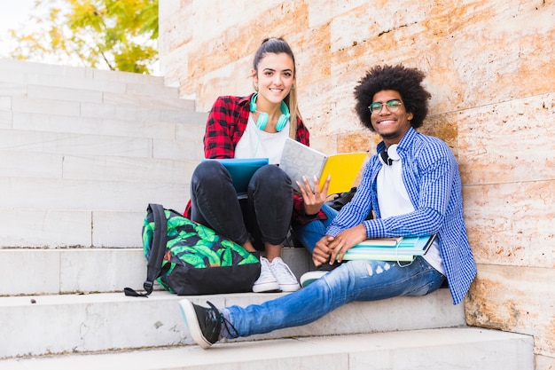 Szczęśliwi męscy i żeńscy studenci uniwersytetu siedzi na schody patrzeje kamera