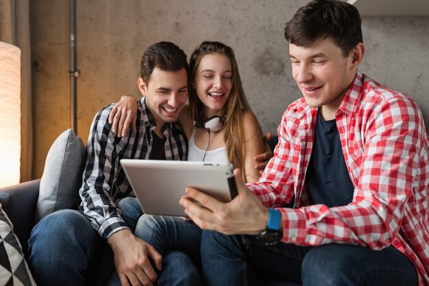 Szczęśliwi młodzi ludzie używający tabletu, uczniowie uczący się, dobrze się bawią, impreza z przyjaciółmi w domu, firma hipster razem, dwóch mężczyzn i jedna kobieta, uśmiechnięta, pozytywna, edukacja online