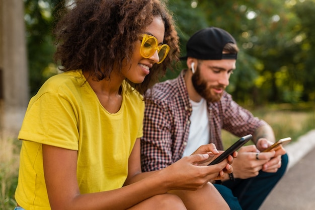 Szczęśliwi młodzi uśmiechnięci przyjaciele siedzi w parku za pomocą smartfonów, mężczyzna i kobieta, wspólna zabawa
