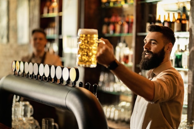 Bezpłatne zdjęcie szczęśliwy barista trzymający szklankę piwa z beczki podczas pracy przy ladzie barowej