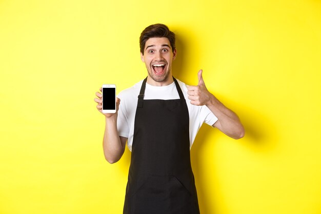 Szczęśliwy barista w czarnym fartuchu pokazuje ekran smartfona, zrób kciuk, polecając aplikację kawiarnianą, stojąc na żółtym tle.