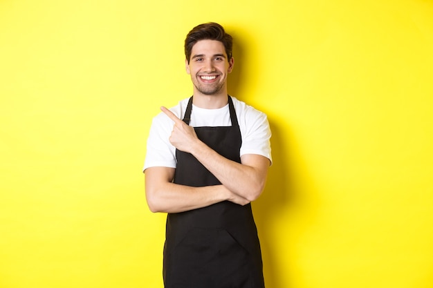 Szczęśliwy barista wskazujący palec w lewo i uśmiechnięty ubrany w czarny fartuch, stojący na tle żółtego b...