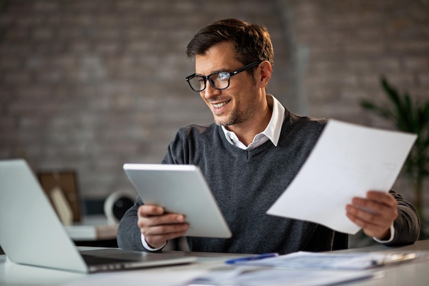 Bezpłatne zdjęcie szczęśliwy biznesmen używający touchpada i laptopa podczas pracy nad raportami biznesowymi w biurze