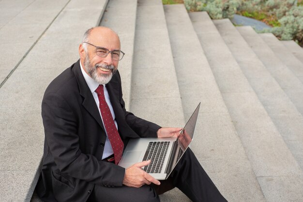 Szczęśliwy biznesmen w eyeglasses używać laptop na ulicie