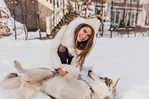 Szczęśliwy czas zimowy niesamowitej uśmiechniętej kobiety z psem husky w śniegu. Urocza młoda kobieta z długimi włosami brunetki, zabawy ze zwierzakiem na ulicy pełnej śniegu. Jasne, prawdziwe emocje.