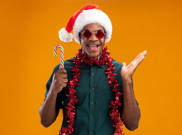 Bezpłatny plik Zdjęcie szczęśliwy człowiek afroamerykanin w santa hat z girlandą w okularach trzyma laskę cukierków, uśmiechając się radośnie z podniesioną ręką stojącą nad pomarańczową ścianą