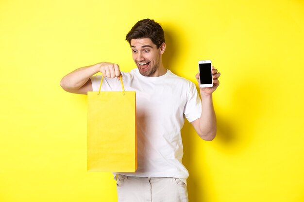 Szczęśliwy człowiek patrząc na torbę na zakupy i pokazując ekran telefonu komórkowego. Koncepcja bankowości internetowej i pieniądza