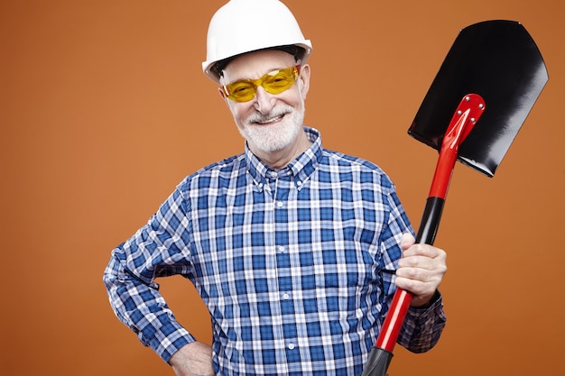Szczęśliwy, energiczny starszy mężczyzna z łopatą do kopania, podnoszenia i przenoszenia materiałów sypkich z szarym zarostem, z pozytywnym, pewnym siebie uśmiechem. Sprzęt budowlany, narzędzia i instrumenty
