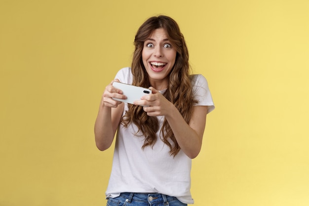 Szczęśliwy entuzjastyczny podekscytowany ładna figlarna dziewczyna wygrywająca niesamowitą grę smartfona zachwycony graniem gapienie się aparat zaskoczony triumfujący uśmiechnięty szczęśliwie trzymający telefon komórkowy poziome żółte tło
