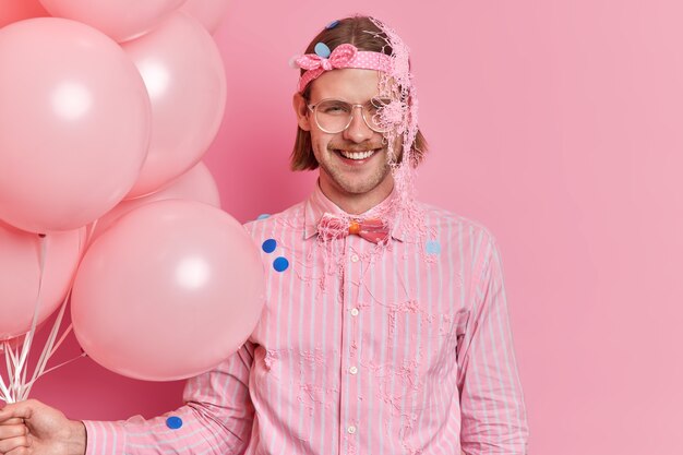 Szczęśliwy europejski dorosły mężczyzna nosi opaskę i koszulę w paski z muszką posmarowaną wężowym sprayem cieszy się przyjęciem z balonów izolowanych na różowej ścianie