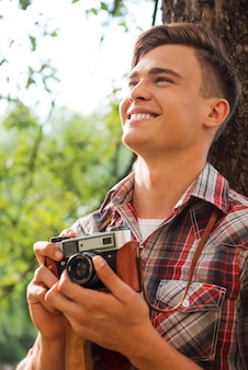 Szczęśliwy fotograf. przystojny młody mężczyzna trzyma starą kamerę i uśmiecha się, opierając się o drzewo