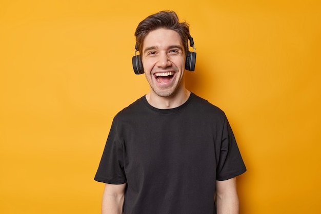 Szczęśliwy hipster facet śmieje się radośnie i ma optymistyczny nastrój słucha ulubionej muzyki przez słuchawki stereo nosi swobodną czarną koszulkę wyizolowaną na żywym żółtym tle Ludzie hobby i koncepcja stylu życia