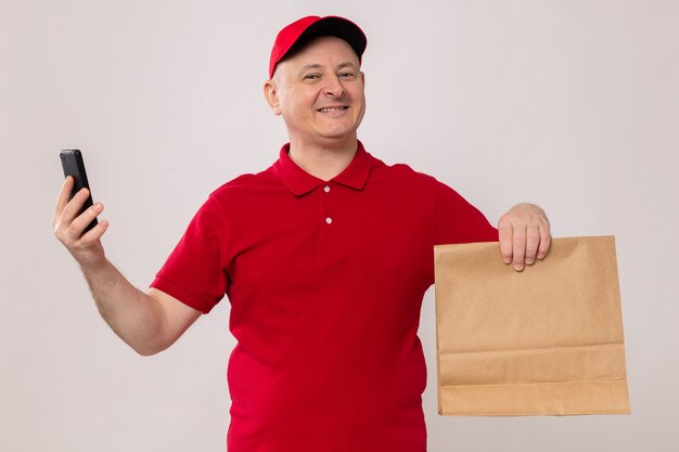 Szczęśliwy i podekscytowany doręczyciel w czerwonym mundurze i czapce, trzymający papierową paczkę i smartfona, patrzący w kamerę, uśmiechający się pewnie stojący na białym tle