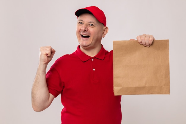 Szczęśliwy i podekscytowany doręczyciel w czerwonym mundurze i czapce trzymający papierowy pakiet patrząc na kamerę uśmiechający się radośnie zaciskający pięść stojący na białym tle