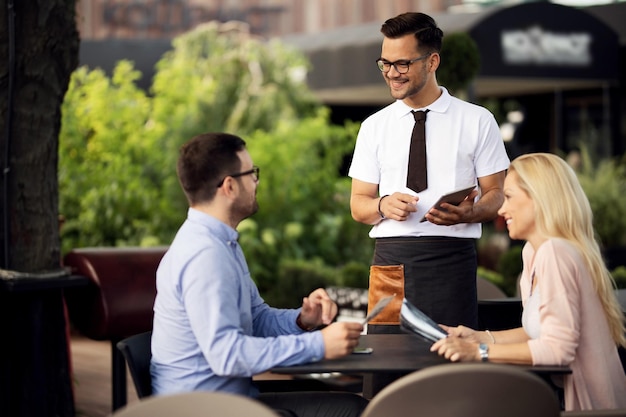 Szczęśliwy kelner korzystający z cyfrowego tabletu podczas rozmowy z parą i przyjmowania od nich zamówienia w restauracji