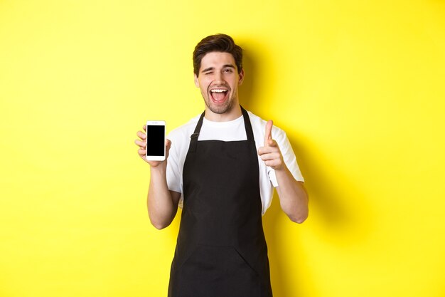 Szczęśliwy kelner pokazujący ekran telefonu komórkowego i kciuk w górę, polecający aplikację kawiarni-restauracji, stojący na żółtym tle.