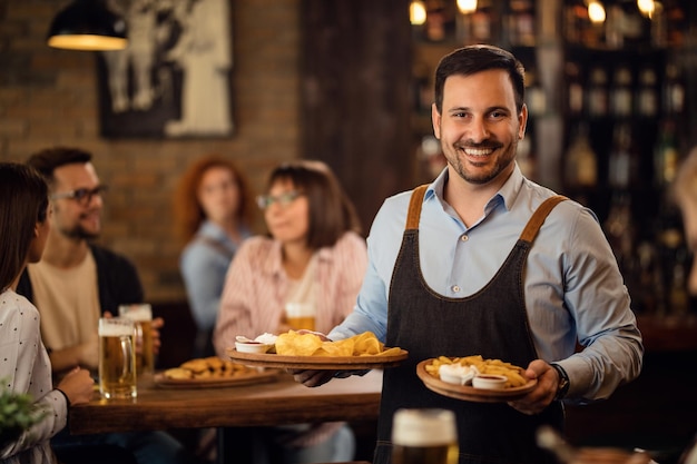 Szczęśliwy kelner trzymający talerze z jedzeniem i patrzący w kamerę podczas obsługi gości w restauracji