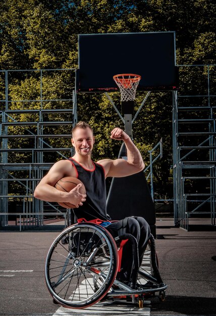 Szczęśliwy koszykarz w pozie na wózku inwalidzkim z piłką na otwartym boisku.