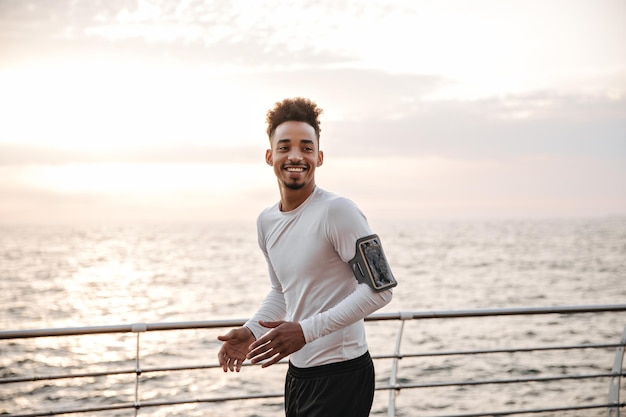Szczęśliwy kręcony ciemnoskóry mężczyzna w białej koszulce i czarnych spodenkach biegnących w pobliżu morza Młody facet ćwiczący i uśmiechający się na zewnątrz