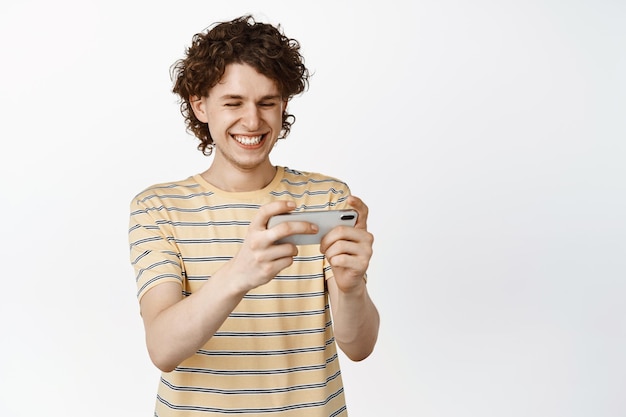 Szczęśliwy kręcony facet grający w grę wideo na telefonie komórkowym, oglądający wideo na ekranie smartfona i uśmiechający się, stojąc na białym tle