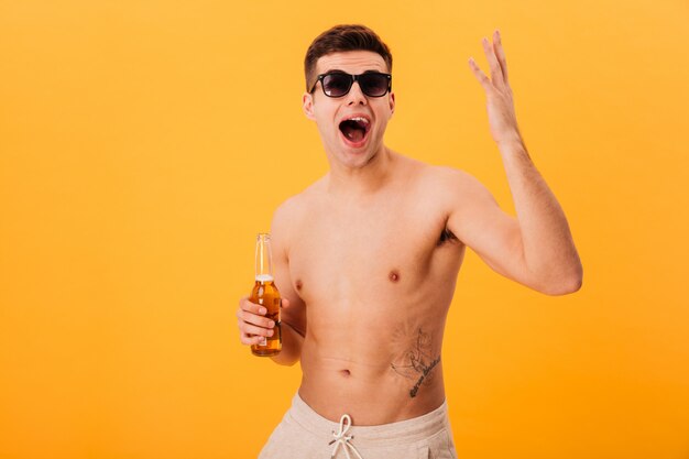 Szczęśliwy krzyczący nagi mężczyzna w szortach i okularach przeciwsłonecznych, trzymając butelkę piwa na żółtym