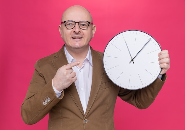 Bezpłatne zdjęcie szczęśliwy łysy mężczyzna w średnim wieku w garniturze w okularach trzymający zegar ścienny wskazujący na niego palcem wskazującym uśmiechnięty wesoło stojąc nad różową ścianą
