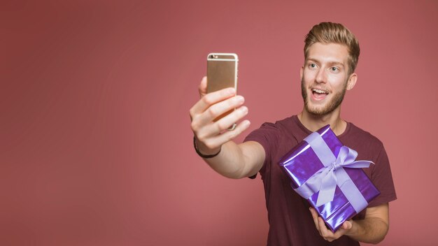 Szczęśliwy mężczyzna bierze selfie z telefonu komórkowego mienia prezenta pudełkiem przeciw barwionemu tłu