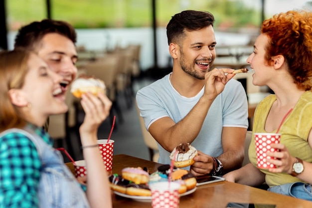Szczęśliwy mężczyzna karmi swoją dziewczynę pączkiem siedząc z przyjaciółmi w kawiarni