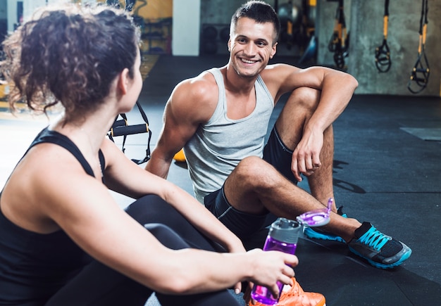 Szczęśliwy mężczyzna obsiadanie na podłogowej patrzeje kobiecie w gym