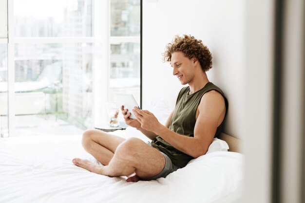 Szczęśliwy mężczyzna siedzi na łóżku w domu przy użyciu komputera typu tablet