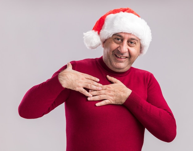 Szczęśliwy mężczyzna w średnim wieku w świątecznym kapeluszu świętego mikołaja, trzymający się za ręce na piersi, czujący wdzięczność uśmiechnięty stojąc nad białą ścianą