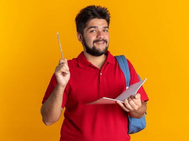 Szczęśliwy młody brodaty student w czerwonej koszulce polo z plecakiem trzymającym notatnik i ołówek patrzący uśmiechnięty radośnie stojący