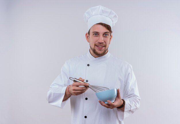 Szczęśliwy młody brodaty szef kuchni ubrany w biały mundur kuchenki i kapelusz trzyma niebieską miskę z łyżką miksera, patrząc na białej ścianie