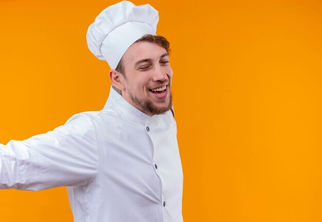 Szczęśliwy młody brodaty szef kuchni w białym mundurze, otwierając szeroko ramiona z zamkniętymi oczami na pomarańczowej ścianie
