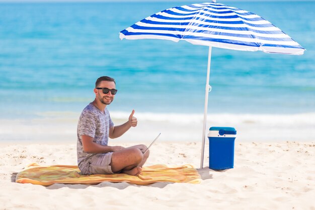 Szczęśliwy młody człowiek na plaży pod parasolem w pobliżu oceanu pracuje na swoim laptopie i pokazuje kciuk w górę