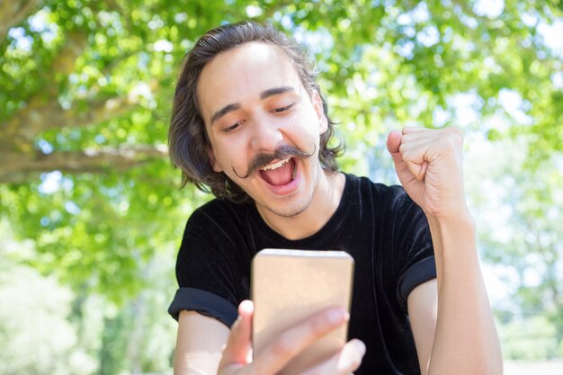 Szczęśliwy młody człowiek patrzeje smartphone w parku.