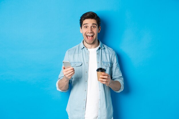 Szczęśliwy młody człowiek pijący kawę i używający telefonu komórkowego, wyglądający na podekscytowanego, stojący na niebieskim tle
