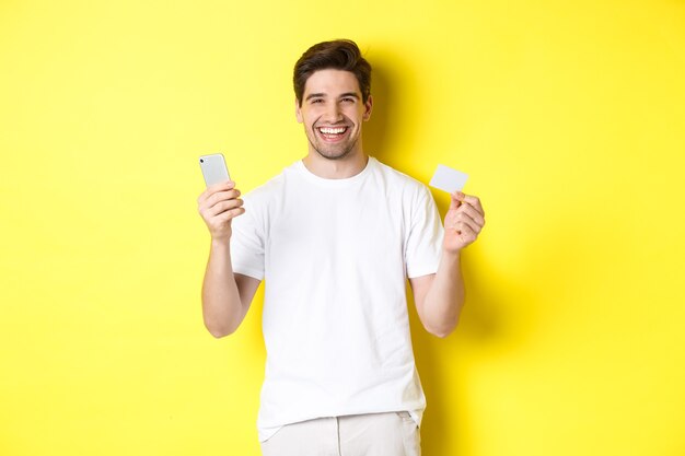 Szczęśliwy młody człowiek robi zakupy online w smartfonie, trzymając kartę kredytową i uśmiechając się, stojąc na żółtym tle