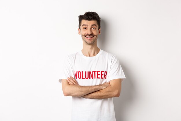 Szczęśliwy młody człowiek w koszulce wolontariusza gotowy do pomocy, uśmiechający się do kamery, skrzyżowane ramiona na klatce piersiowej pewny siebie, białe tło