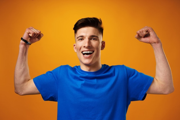 Szczęśliwy młody człowiek w niebieskiej koszulce uśmiechający się z uniesionymi rękami świętujący sukces