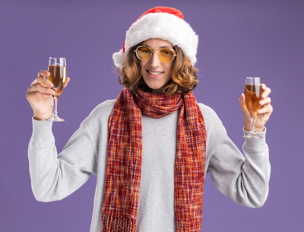 szczęśliwy młody człowiek w świątecznej czapce mikołaja i żółtych okularach z ciepłym szalikiem na szyi trzymający kieliszki szampana patrząc w kamerę uśmiechnięty wesoło stojąc na fioletowym tle