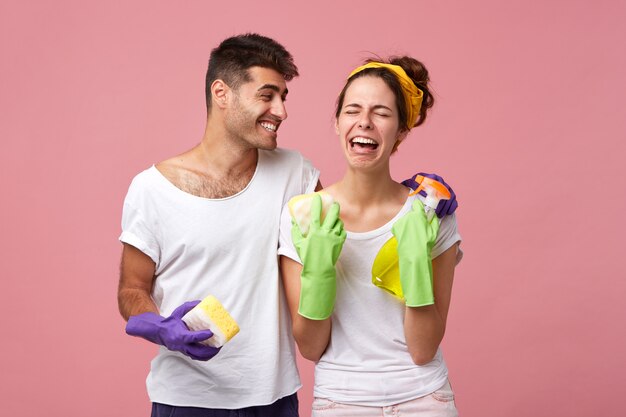 Szczęśliwy młody mężczyzna pocieszający zdesperowaną zestresowaną kobietę w ochronnych rękawiczkach, która nie ma ochoty zmywać naczyń. Przystojny pozytywny mężczyzna śmiejący się ze swojej smutnej, płaczącej dziewczyny, która nienawidzi sprzątania