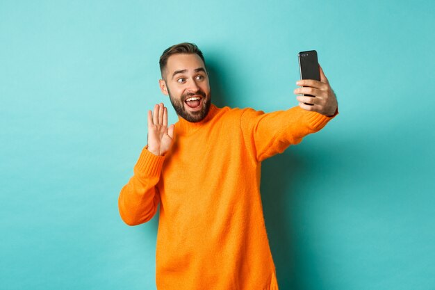 Szczęśliwy młody mężczyzna prowadzi rozmowy wideo, rozmawia przez Internet z telefonem komórkowym, wita się z aparatem smartfona i macha ręką, stojąc nad jasnoniebieską ścianą.