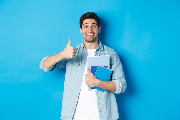 Szczęśliwy młody model płci męskiej trzymający zeszyty i pokazujący zadowolony kciuk w górę, uśmiechnięty i polecający kursy, stojący na niebieskim tle